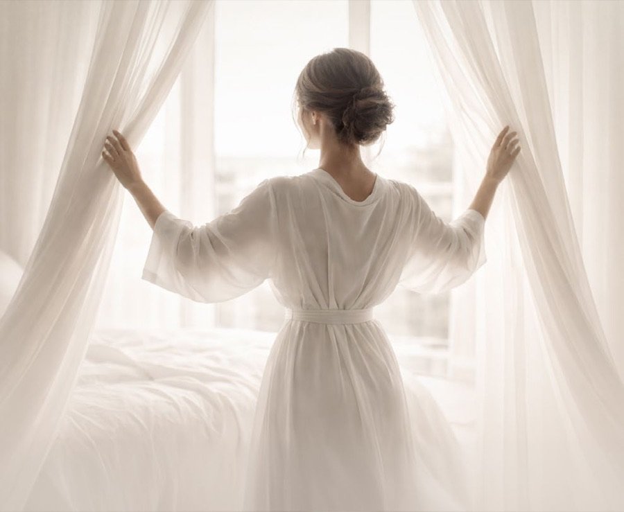 A woman drawing back sheer curtains onto morning light